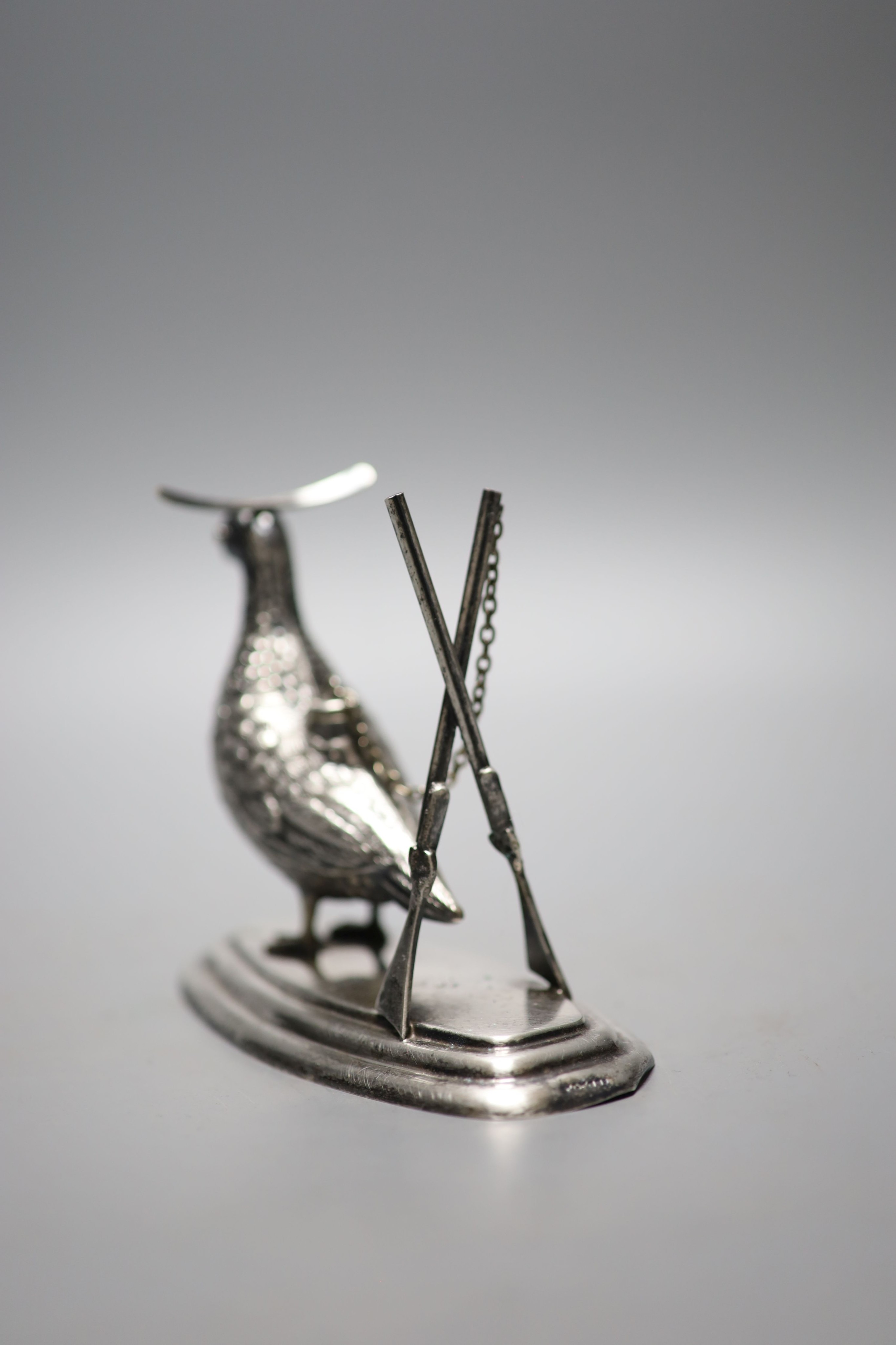 A continental novelty white metal brand balloon warmer, modelled and a game bird with crossed rifles, on a shaped oval base, 13.1cm, indistinct stamp.